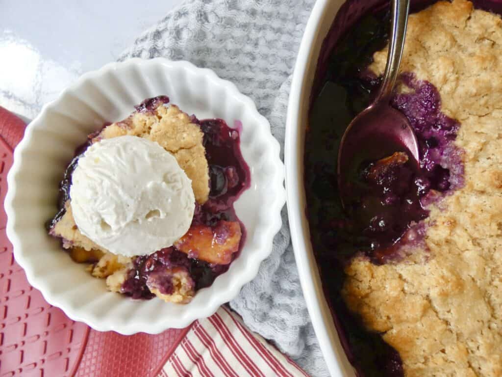 bowl of healthier blueberry peach cobbler with vanilla ice cream next to baking dish